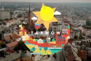 Kolorowe logo "Tarnów Hot Spot" na tle miasta nakręconego z lotu ptaka. Logo jest wzorowane na herbie miasta – leliwie. Leliwa to księżyc pod sześcioramienną gwiazdą. Gwiazdą z herbu jest słońce a księżycem - charakterystyczne budynki i zieleń Tarnowa.